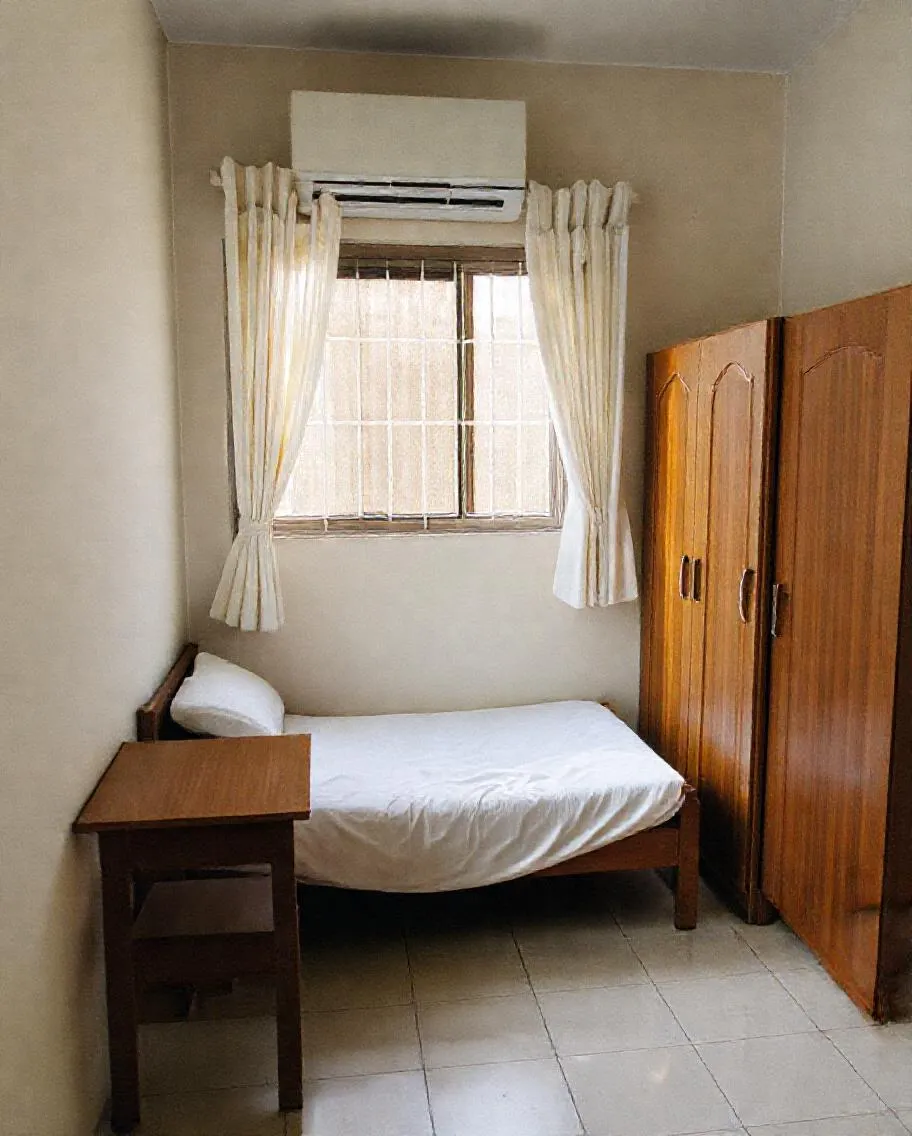 Kamar Wisma Melati: kasur berseprai putih, meja belajar di bawah jendela bertirai tipis, AC di dinding