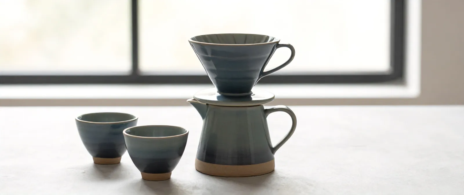 The Kiln set: a ribbed stoneware dripper on its carafe, with two handleless cups, in a slate glaze that pools darker at the foot