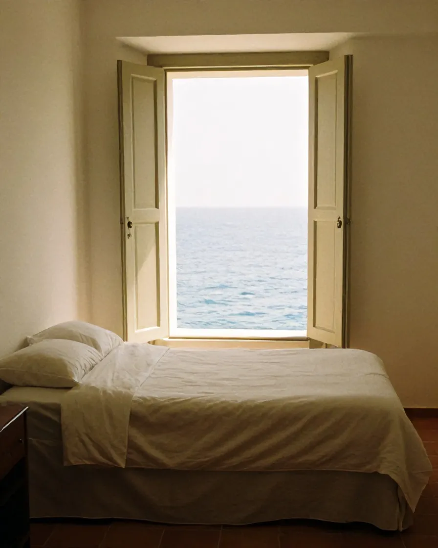 A guest room with linen bed and open shutters onto the sea