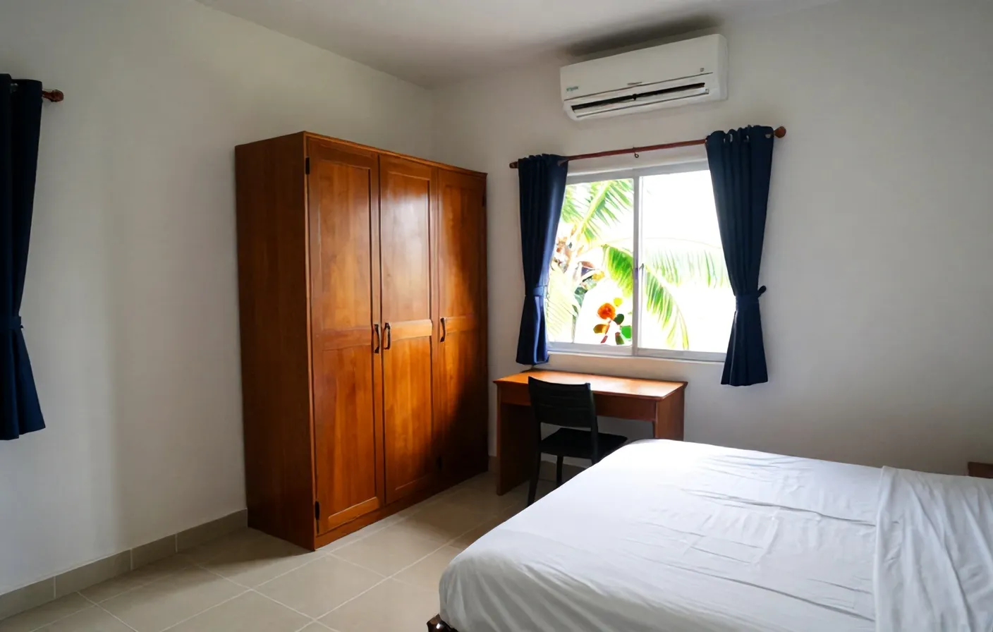 Kamar kos dengan kasur putih, lemari kayu, meja belajar di bawah jendela teralis, dan AC