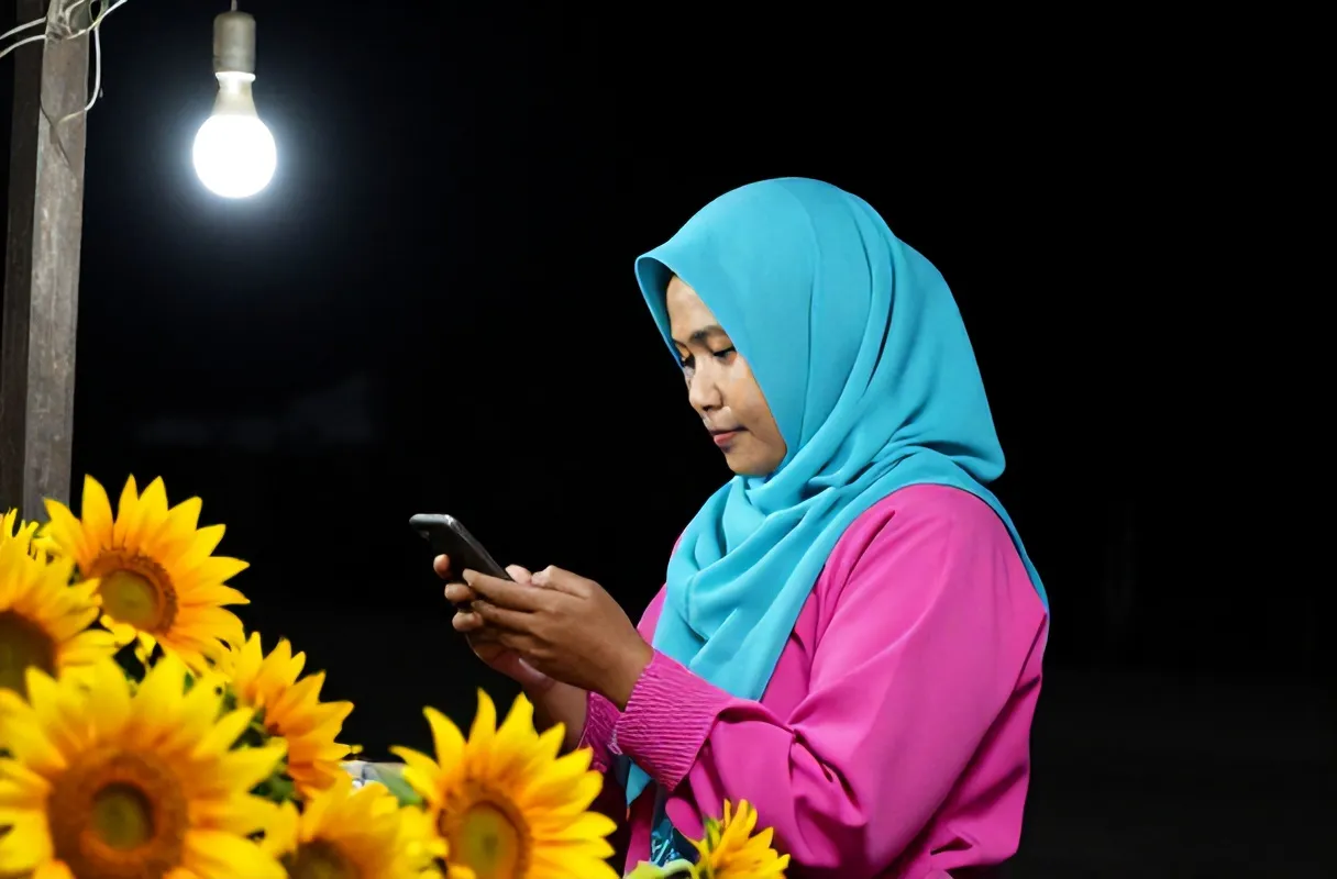 Pemilik warung berdaster menghitung penjualan di HP malam hari di bawah satu bohlam