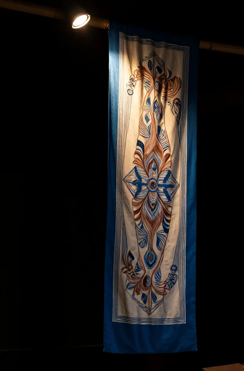Selembar batik tulis indigo dan krem tergantung di bambu, disorot satu lampu di ruang gelap