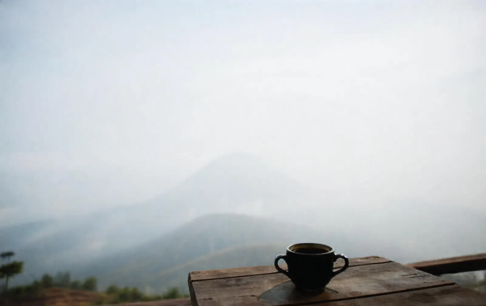 Cangkir kopi hitam di meja kayu teras dengan lereng Merapi berkabut di belakangnya