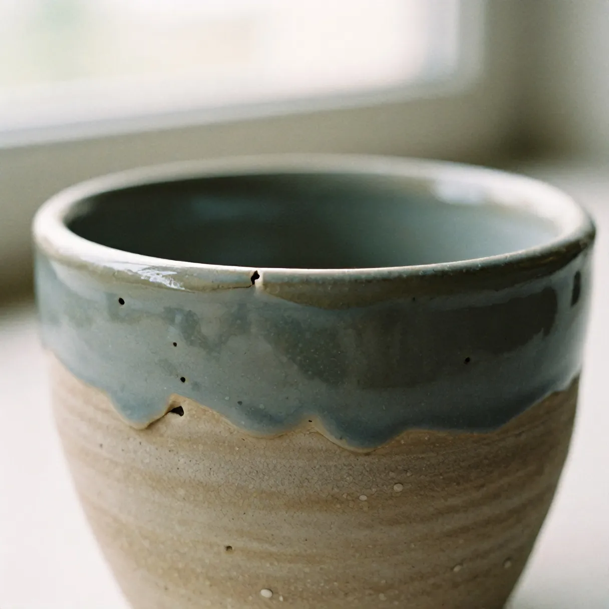 Sage glaze breaking pale over a cup rim, with drips and pinholes
