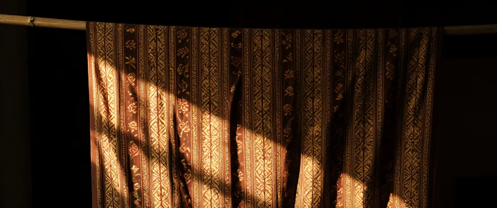 Selembar batik tulis soga cokelat dan krem tergantung di bambu, disinari seberkas cahaya jendela di ruang gelap