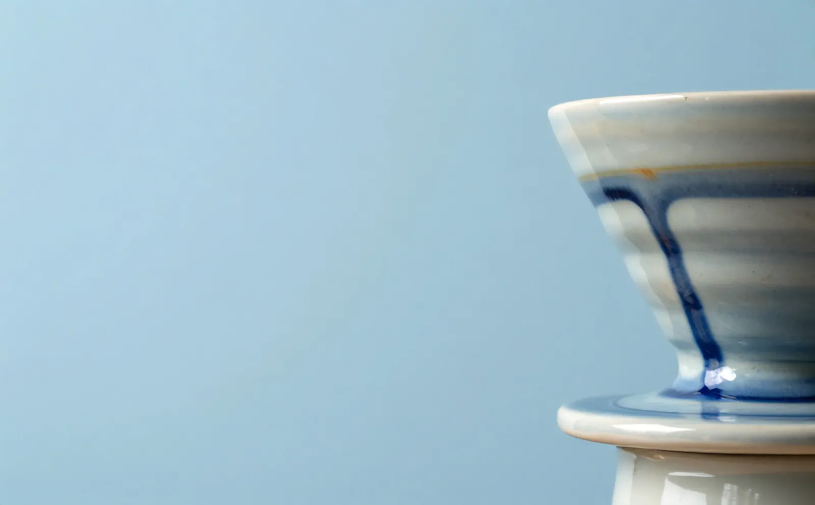 A Kiln pour-over dripper in cream glaze, blue drips running down its side, seen close on a sky-blue backdrop