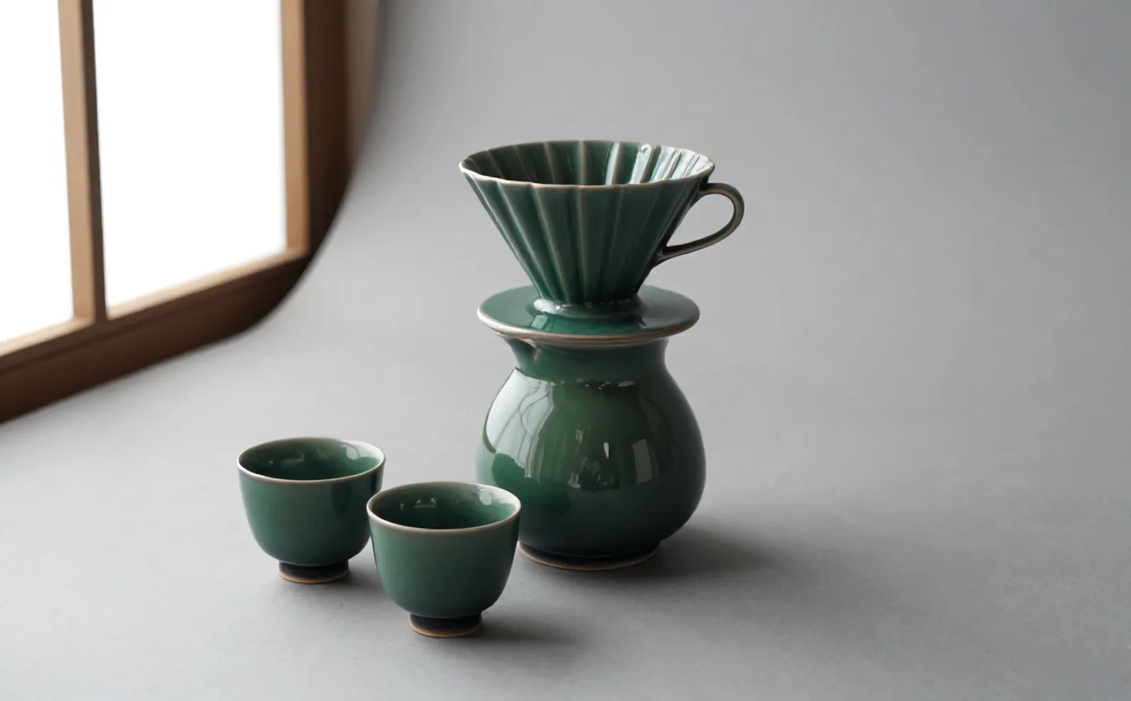 The Kiln set on a grey studio floor: a green-glazed dripper on its carafe, with two small cups beside it