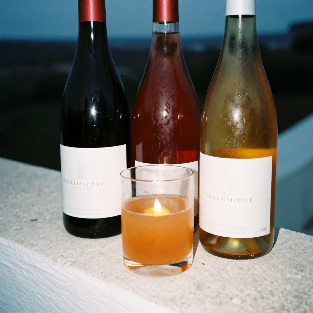 Three natural wine bottles and a tumbler of cloudy orange wine on a whitewashed stone ledge