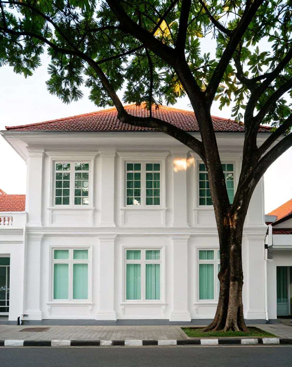Rumah kolonial putih berjendela tinggi di Jl. Raya Darmo 118, di bawah pohon trembesi besar, sore hari