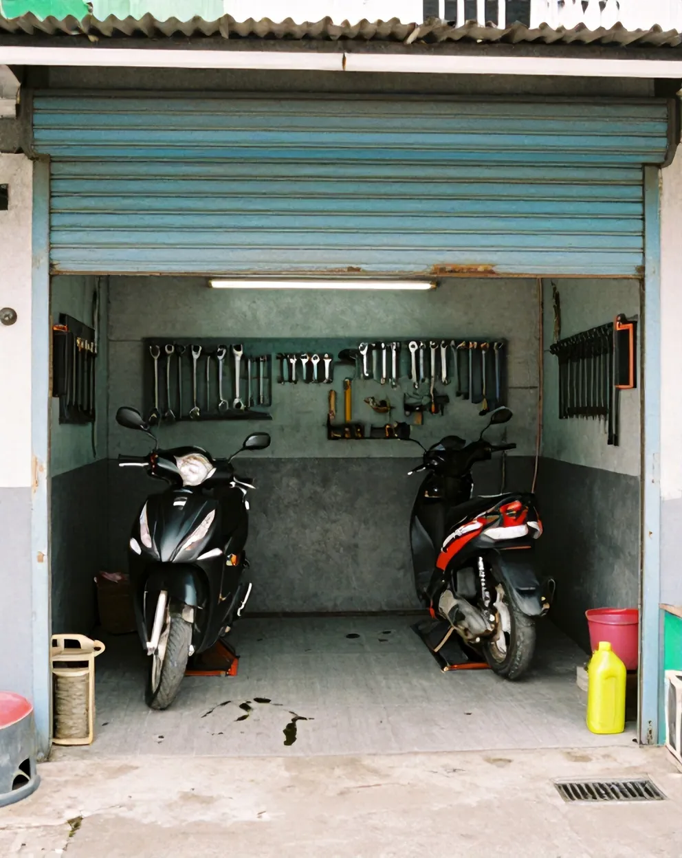 Bagian depan bengkel Jaya Abadi dengan rolling door terbuka, satu skuter di atas lift dan satu lagi menunggu